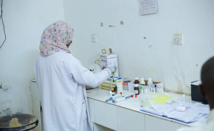 A female health worker in a white coat and flowery headscarf holds a blood sample, facing the wall inside a clinic