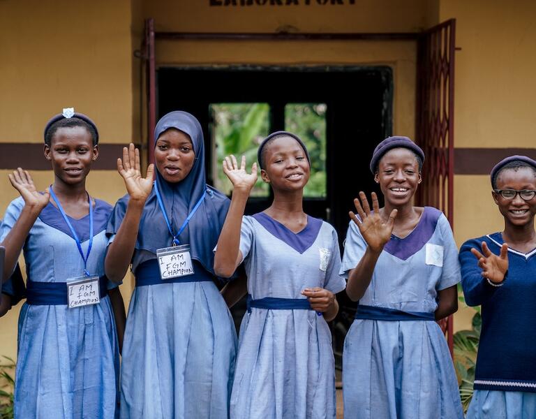 Five girls wearing blue school dresses wave there hands and smile towards.